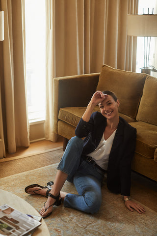 A smiling woman is sitting on the floor of a living room with a velvet couch and a marble coffee table. She is wearing jeans, black sandals, and a navy blazer over Field Trip's reversible flutter sleeve in white worn with the V neck side in front.
